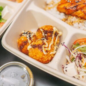 Inicio Close-up of a bento box with grilled salmon, rice, salad, and croquettes.