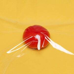 A fresh tomato wrapped in clear plastic wrap on a vibrant yellow background, highlighting freshness and packaging.